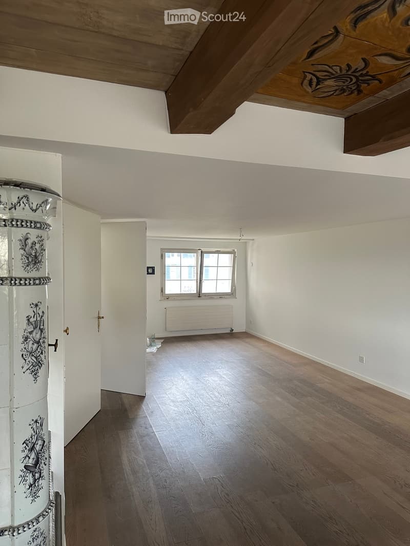 Empty room with wooden flooring, white walls, and a window with white frame. Exposed wooden beams on the ceiling.