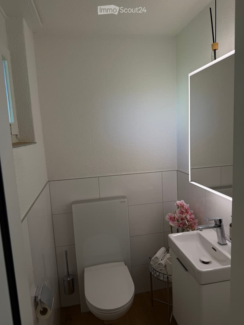 Modern bathroom with a toilet, sink, mirror, and decorative flowers. The room features white tiled walls and a wooden floor.
