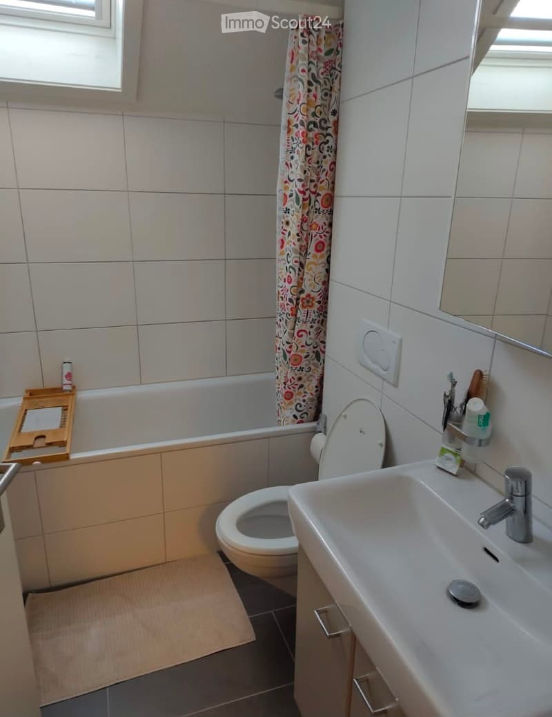 Bathroom with white tiled walls, a toilet, a sink with a faucet, a bathtub with a colorful shower curtain, and a small wooden tray on the edge of the tub.