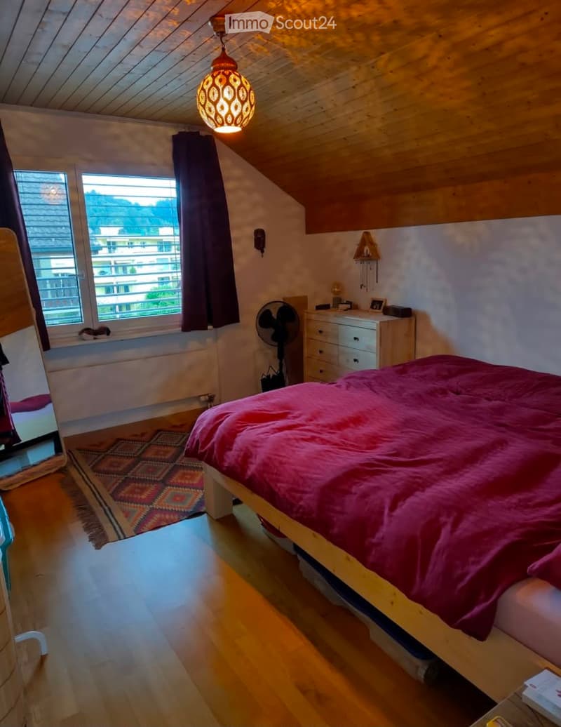 Cozy attic bedroom with wooden walls and ceiling, large window with view, bed with red duvet, fan, nightstand, and mirror.