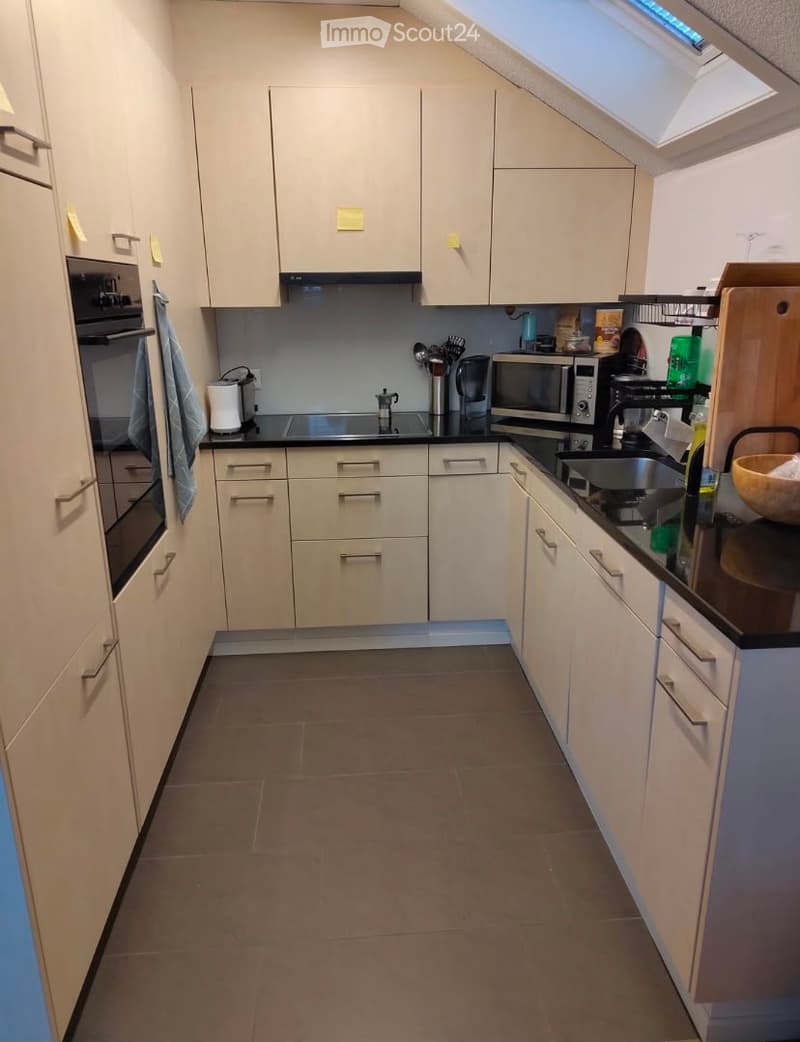 Modern kitchen with light-colored cabinets, stainless steel appliances, and a black countertop. Features include an oven, microwave, refrigerator, sink, and various small appliances.