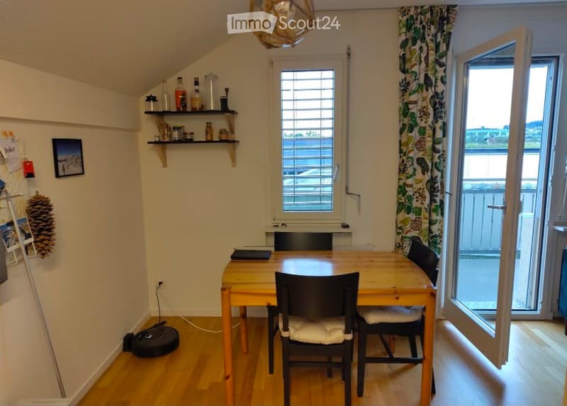 Bright, airy room with wooden flooring, large window with blinds, open door leading to a balcony, wooden dining table with chairs, shelf with various items, and a laptop on the table.