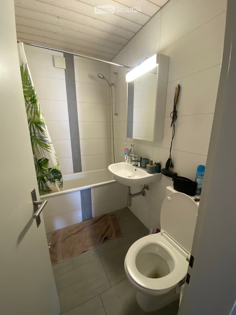 Modern bathroom with white tiles, a bathtub, a sink, a toilet, a mirror, and a showerhead. A towel is placed on the floor, and various toiletries are on the sink counter.