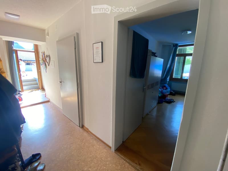 Empty hallway with light-colored walls, wooden flooring, and two open doors leading to other rooms. One door opens to a room with a window and some scattered items, while the other leads to a brightly lit area with outdoor steps visible.