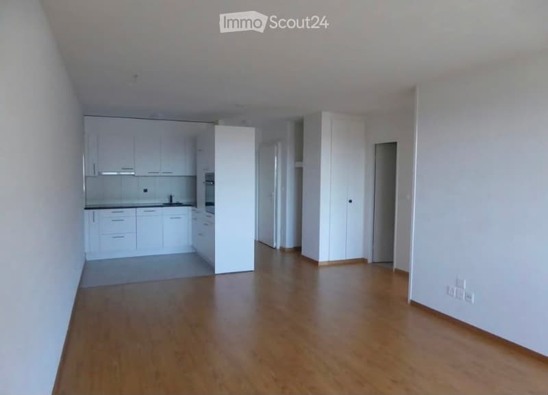 Bright, open-plan living and kitchen area with white cabinets, a sink, and a refrigerator. The floor is wooden, and the walls are white.