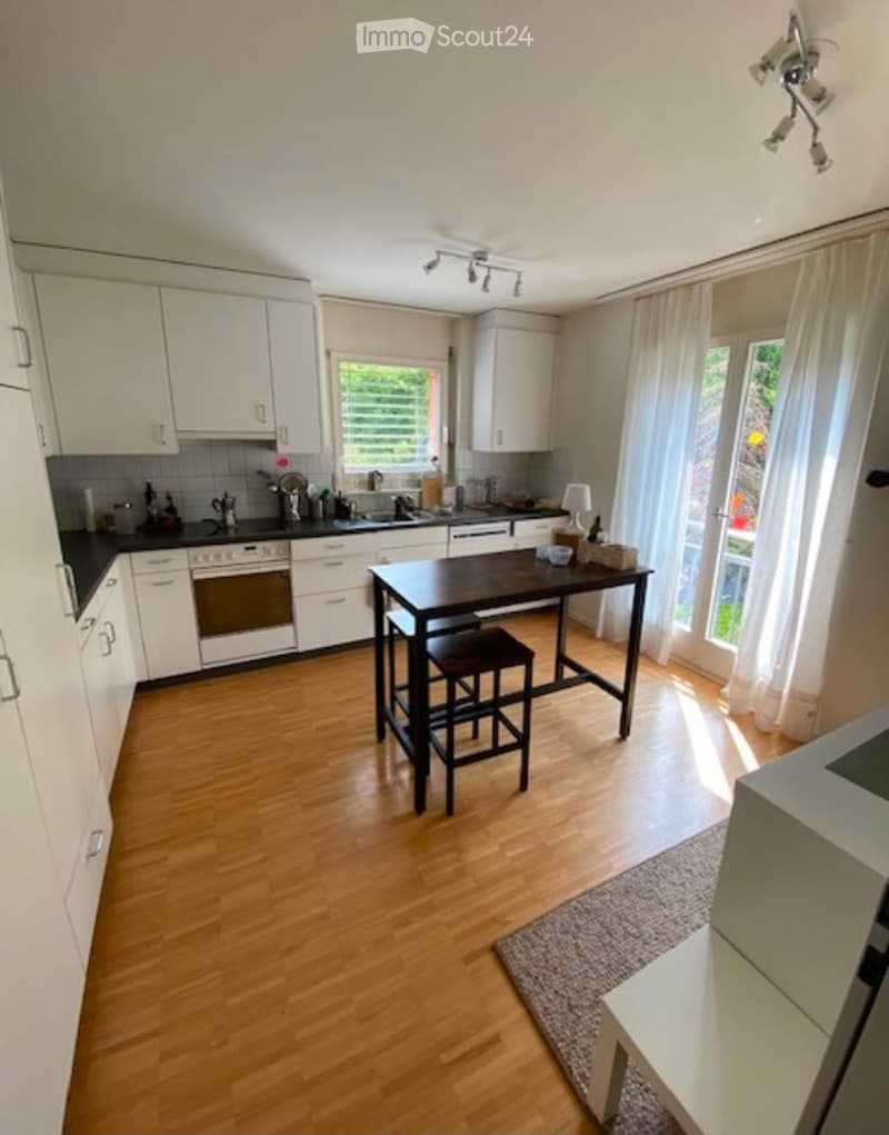 Modern kitchen with white cabinets, black countertops, and wooden flooring. Features a central island table with stools, a built-in oven, and a sink area.