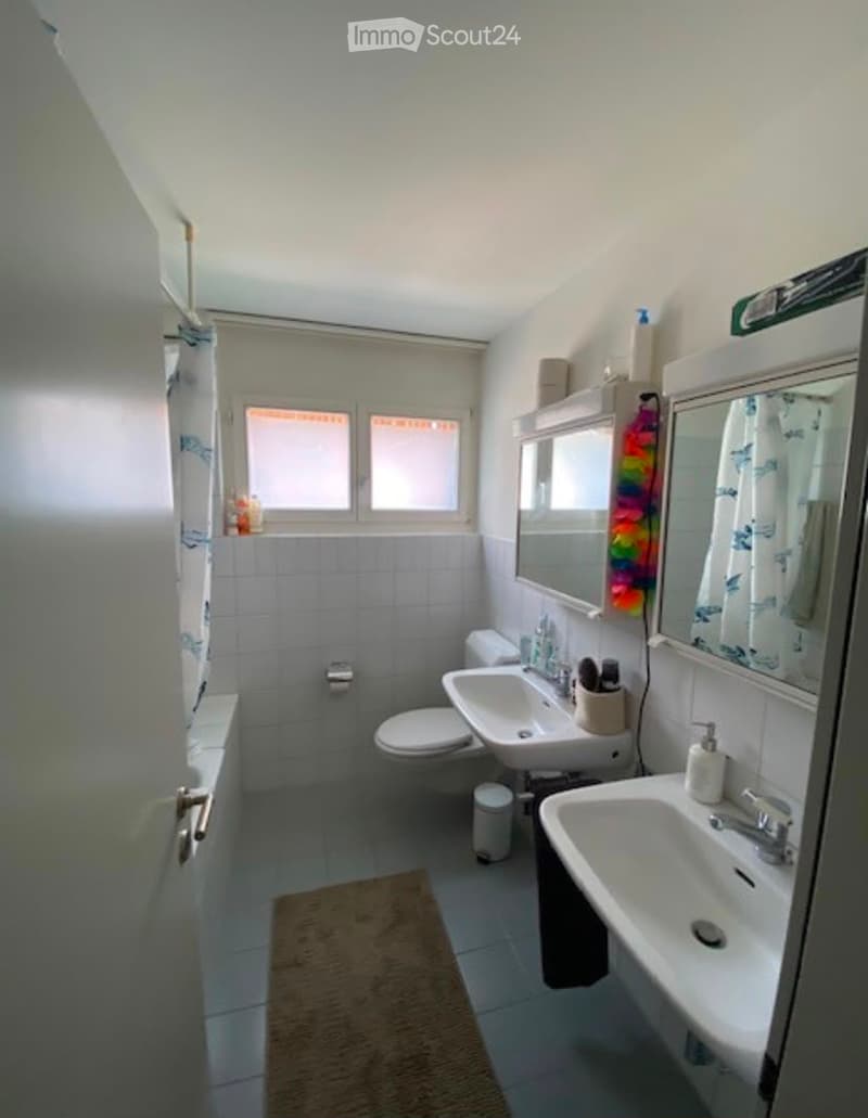 A small, well-lit bathroom with white tiled walls and floor, featuring a toilet, two sinks, a bathtub, a mirror, and a small bathroom rug.
