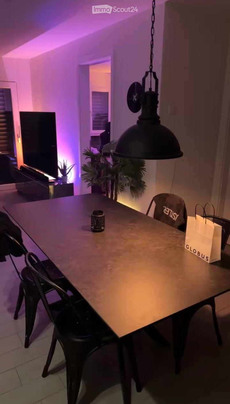 A modern dining area with a large wooden table, black chairs, a pendant light, and a plant in the background.