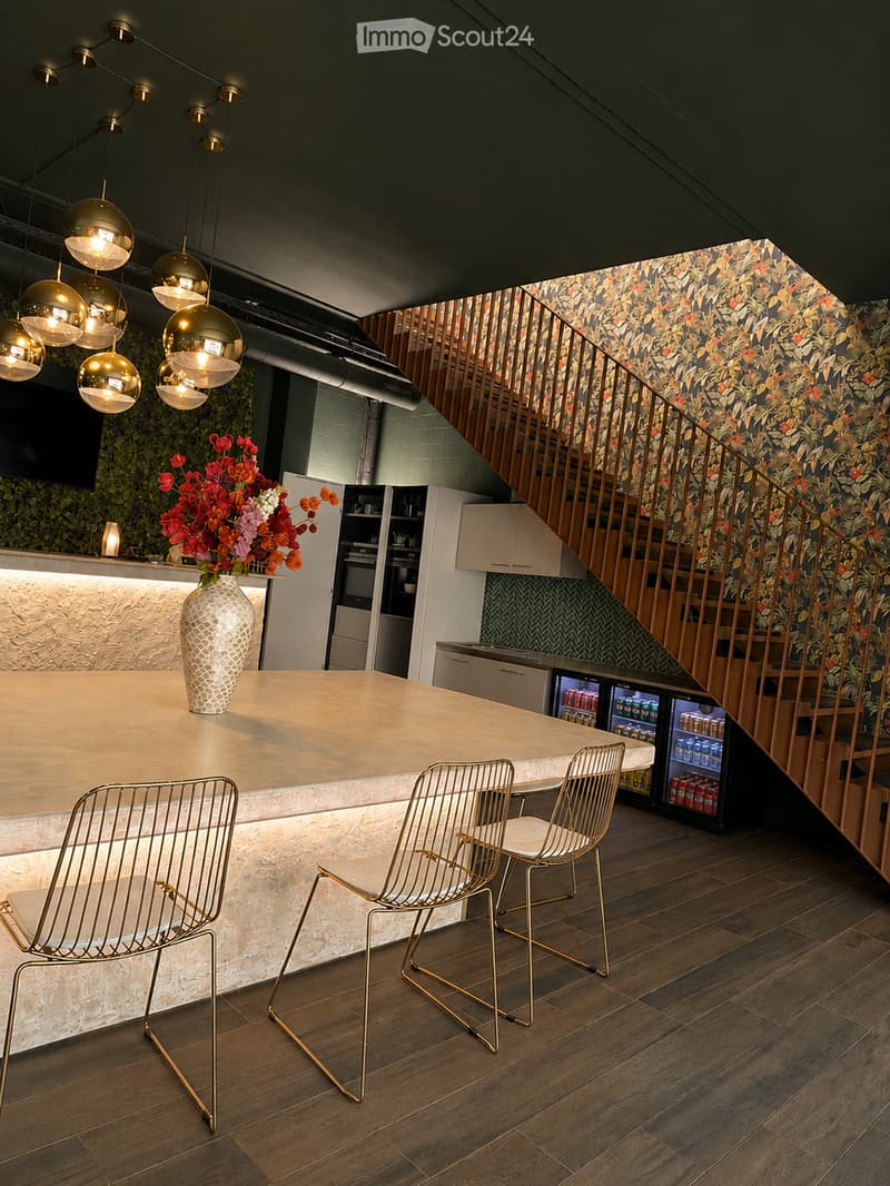Modern bar area with a sleek marble counter, pendant lights, two metal chairs, a vase with flowers, and a small refrigerator. The space features a wooden staircase leading to an upper level.