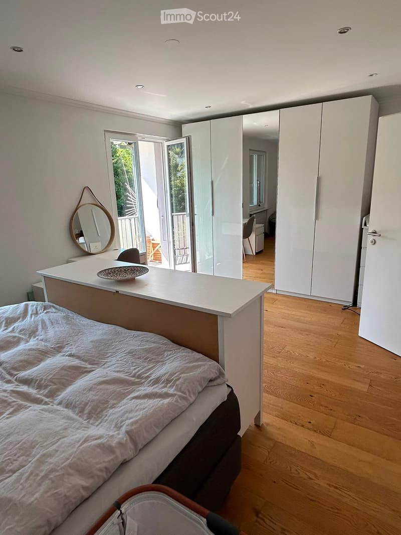Modern bedroom with a large bed, a white table with a decorative bowl, a round mirror on the wall, and a sliding glass door leading to a balcony.