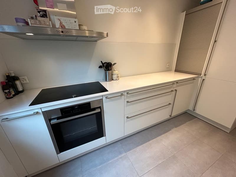 Modern kitchen with white cabinets, an induction stove, an oven, and a kitchen island. The countertops are white, and the floor is tiled.