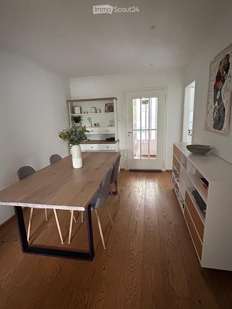 Dining room with wooden floor, modern dining table, chairs, shelves, and a vase with flowers.