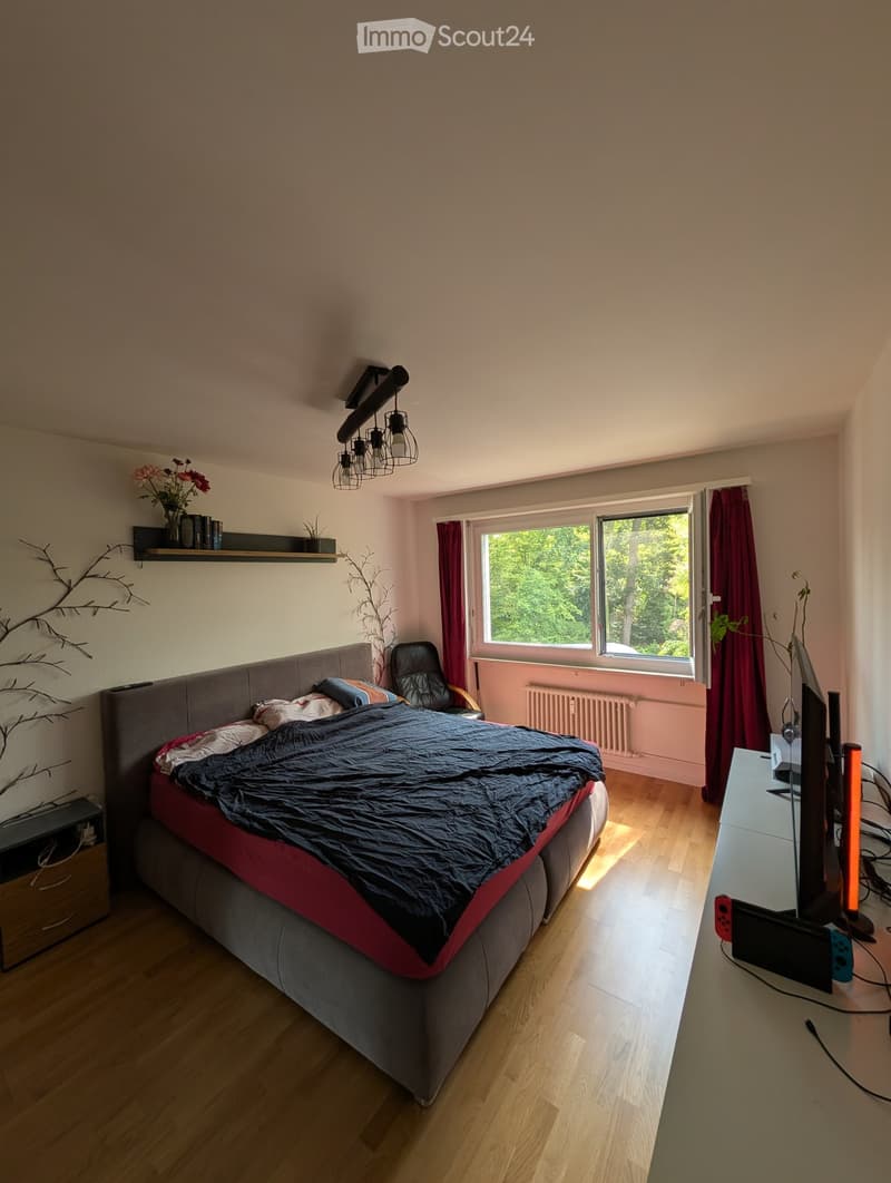 Modern bedroom with a large bed, wooden floor, large window with red curtains, and a desk with a screen and gaming console.
