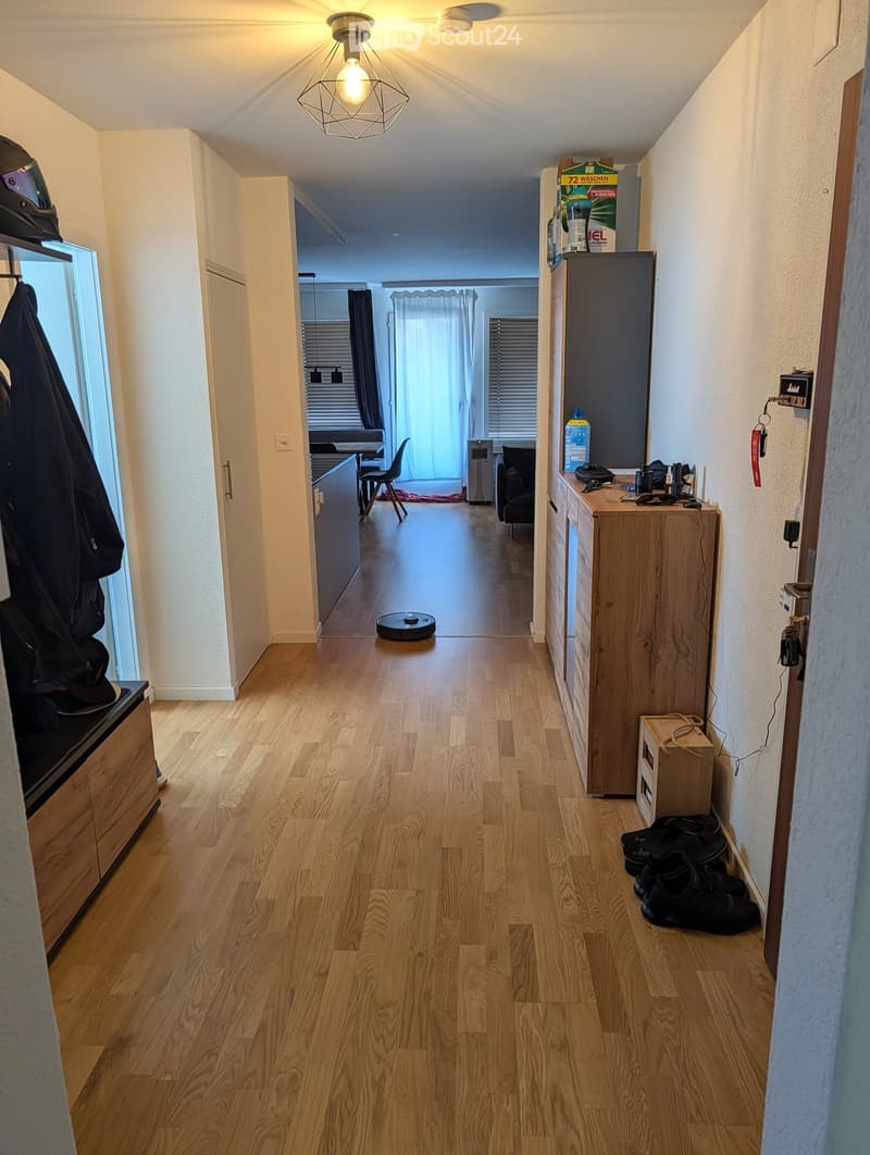 A clean, well-lit hallway with wooden flooring, leading to a room with a dining table and chairs. There are various items like a vacuum cleaner, shoes, and a small table with items on it.