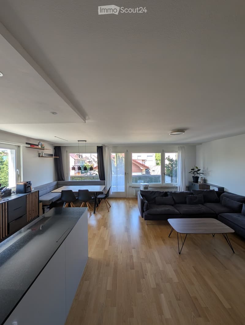 Modern living room with wooden flooring, large windows, a sectional sofa, coffee table, dining area with chairs, and a small kitchen counter.