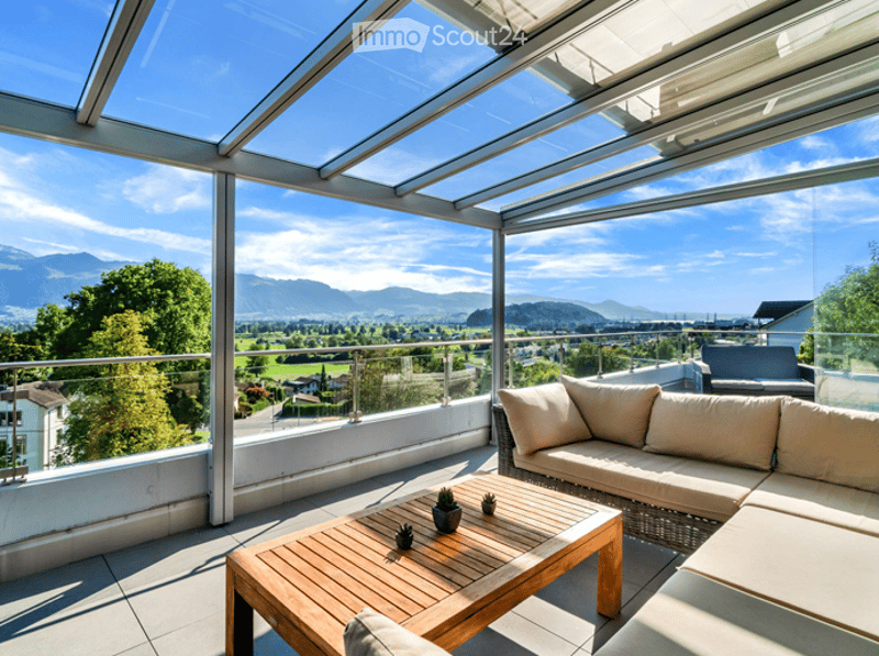 A modern glass-enclosed balcony with a panoramic mountain view, featuring a wooden coffee table, a cream-colored sofa, and small potted plants.