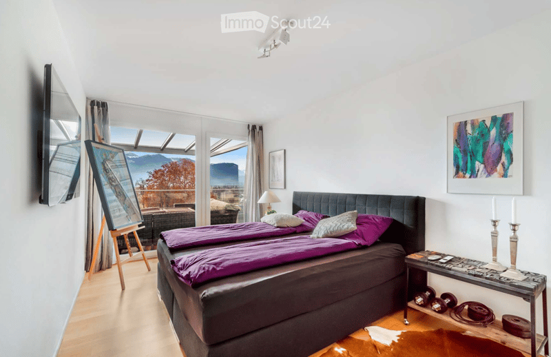 Bright bedroom with a large bed, nightstand, lamp, mirror, picture frame, and a view of the mountains through large windows.