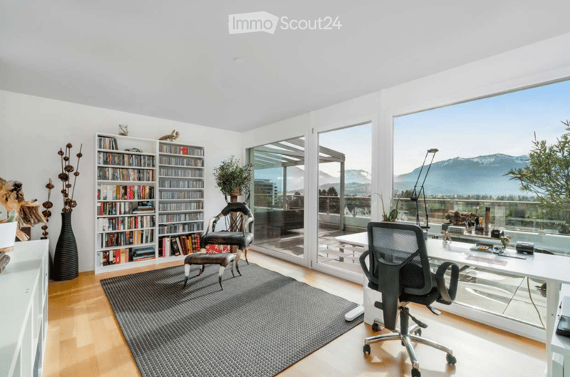 Bright, modern office space with large windows offering a scenic mountain view. Features include a bookshelf, a comfortable chair with a footstool, a desk with an office chair, and decorative vases with plants.