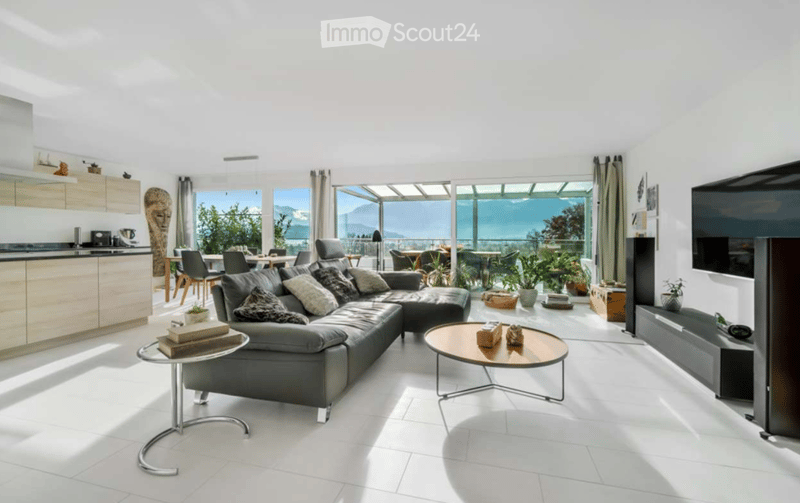Bright, modern living space with white walls, white tiled flooring, and large windows offering a scenic view. Features a grey sectional sofa, coffee tables, a TV stand, kitchen area with cabinets, and a dining area with chairs.