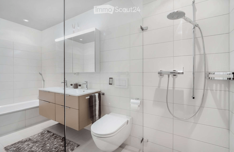 Modern bathroom with white tiles, a glass-enclosed shower, a sink with a cabinet, a toilet, and a bathtub. The space includes a mirror, towel, and light switch.