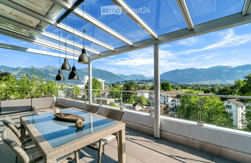 A modern glass-enclosed balcony with a scenic mountain view, featuring a glass table and wicker chairs.