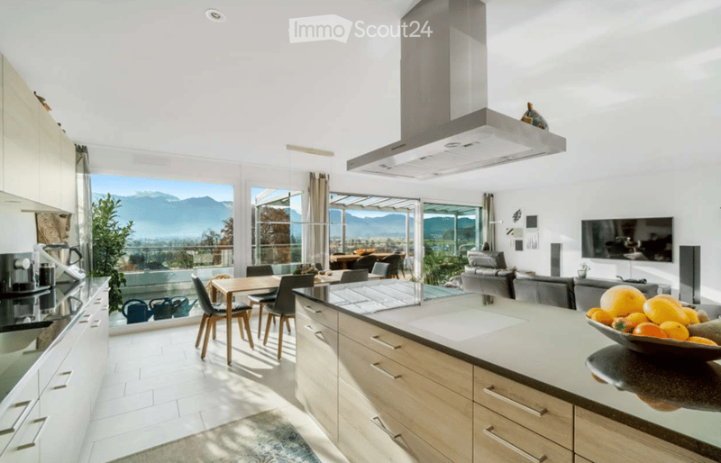 Open-plan kitchen and living area with modern white cabinets, a central island, a dining table with chairs, a sofa, and a television. Large windows offer a scenic mountain view.