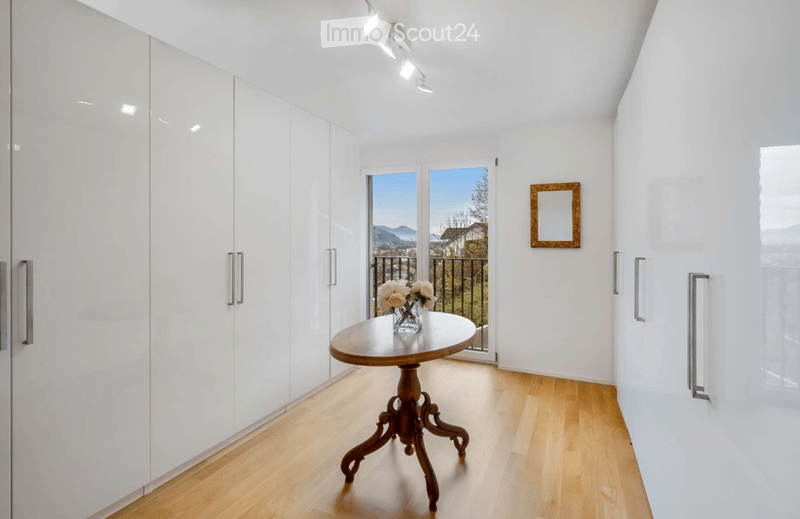 Bright, modern room with white walls, light wooden flooring, built-in white wardrobes, a small round wooden table with a vase of white flowers, and a framed mirror on the wall. Large windows offer a scenic mountain view.