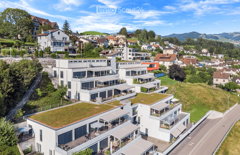 Aerial view of a modern white building with green roofs, surrounded by a scenic town with houses, trees, and mountains in the background.