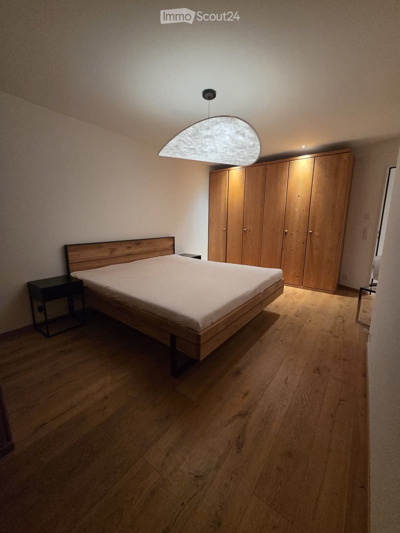 Modern bedroom with wooden floor, large bed with white bedding, built-in wooden wardrobe, and pendant light fixture.