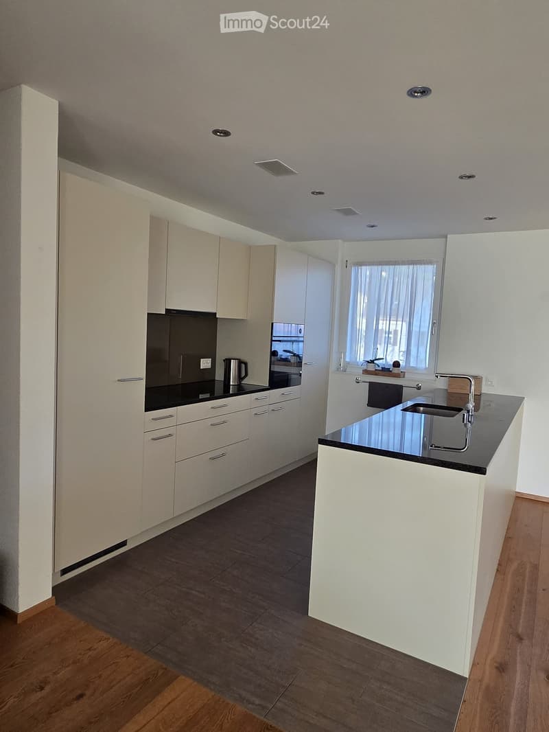 Modern kitchen with white cabinets, black countertops, and stainless steel appliances. Features a central island with a sink and faucet, and a window with a curtain.