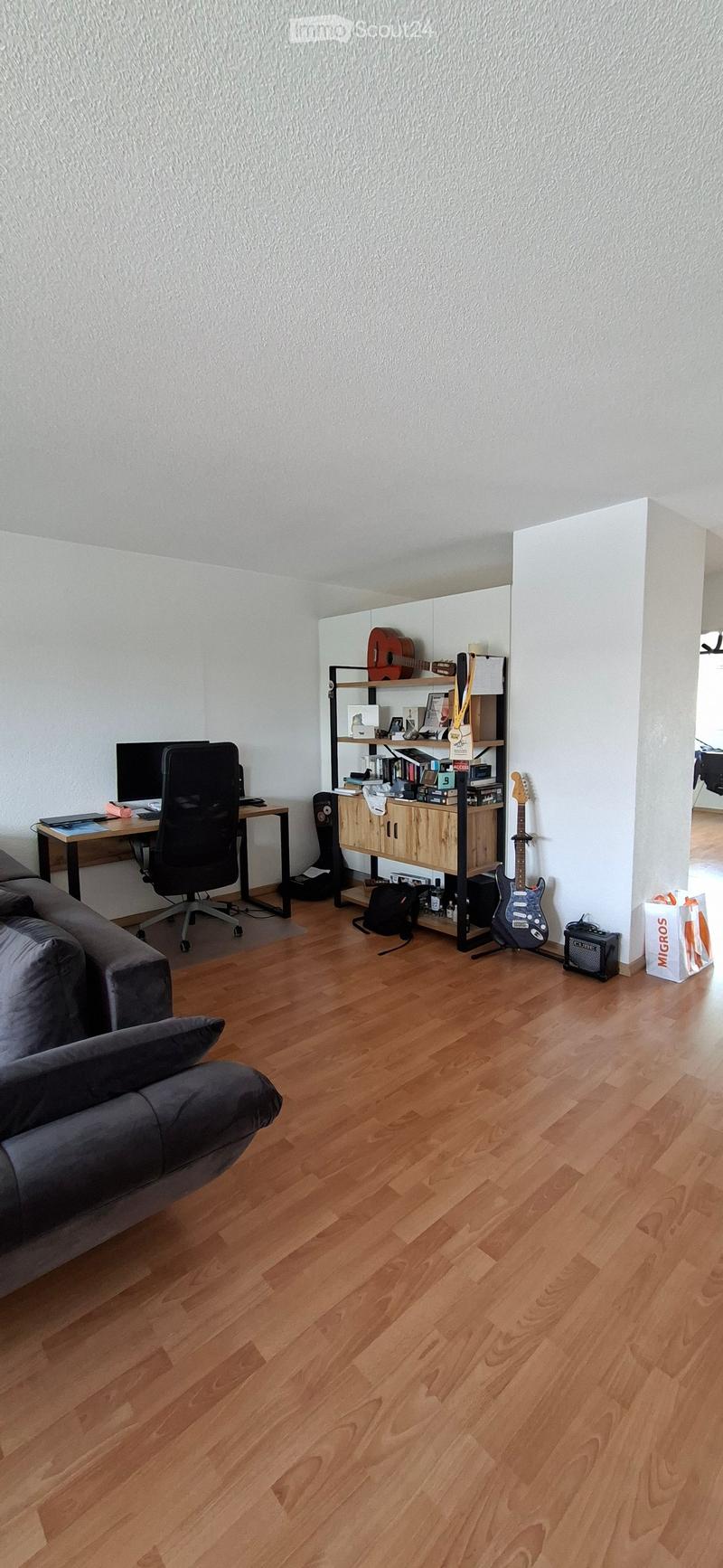 A modern living room with wooden flooring, a grey sofa, a desk with a chair, a guitar and amplifier, and a bookshelf with books and decorative items.