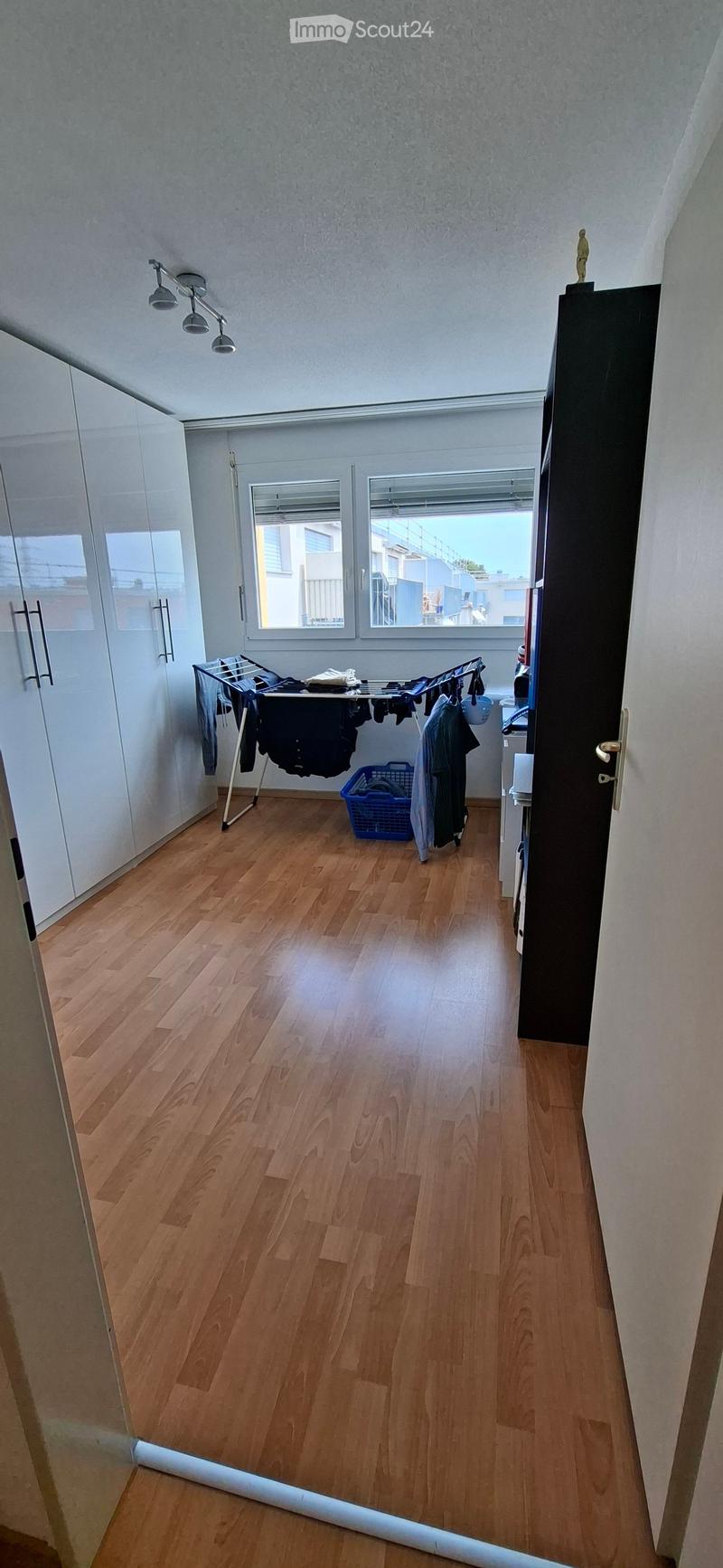 Empty room with wooden flooring, white walls, and large windows. Features a drying rack with clothes, a blue basket, and a partially visible closet.