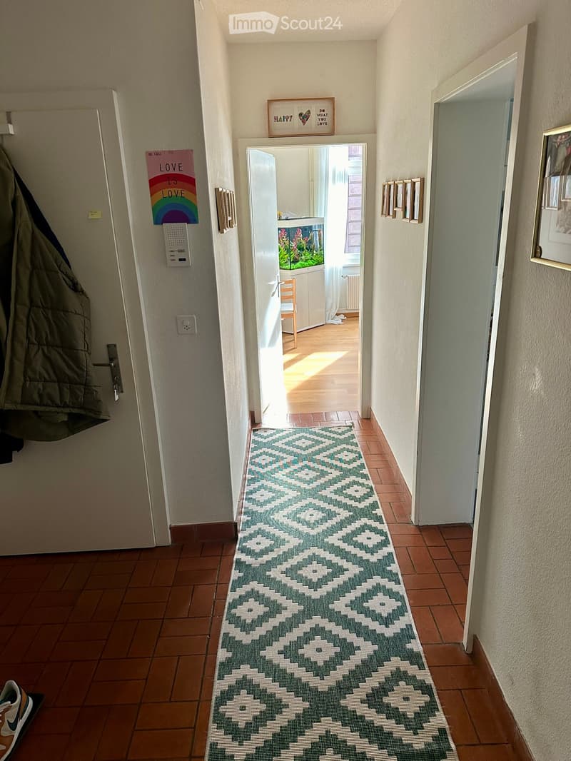 Hallway with tiled floor, patterned runner rug, open door leading to another room, coat hanging on the left, and framed pictures on the right wall.