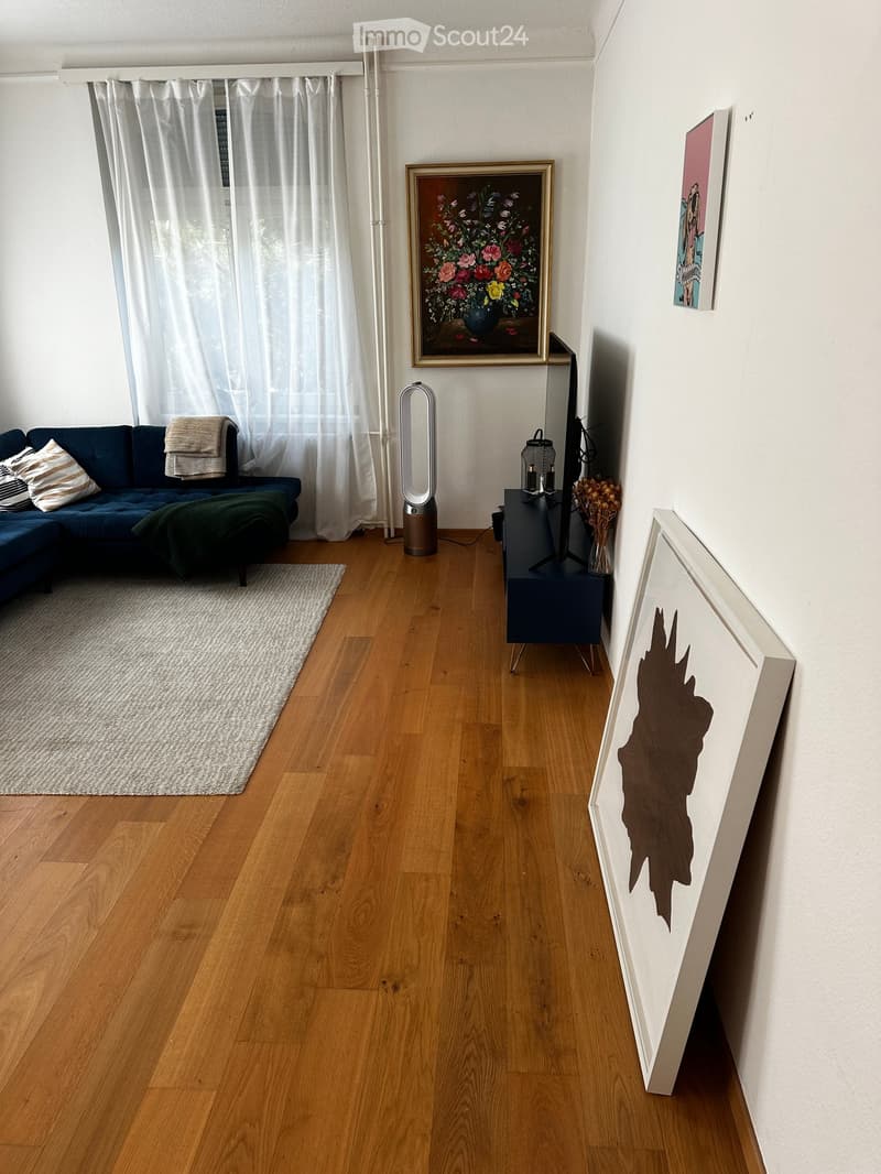 Modern living room with wooden flooring, a blue sofa, a TV stand, a speaker, and several paintings on the walls.
