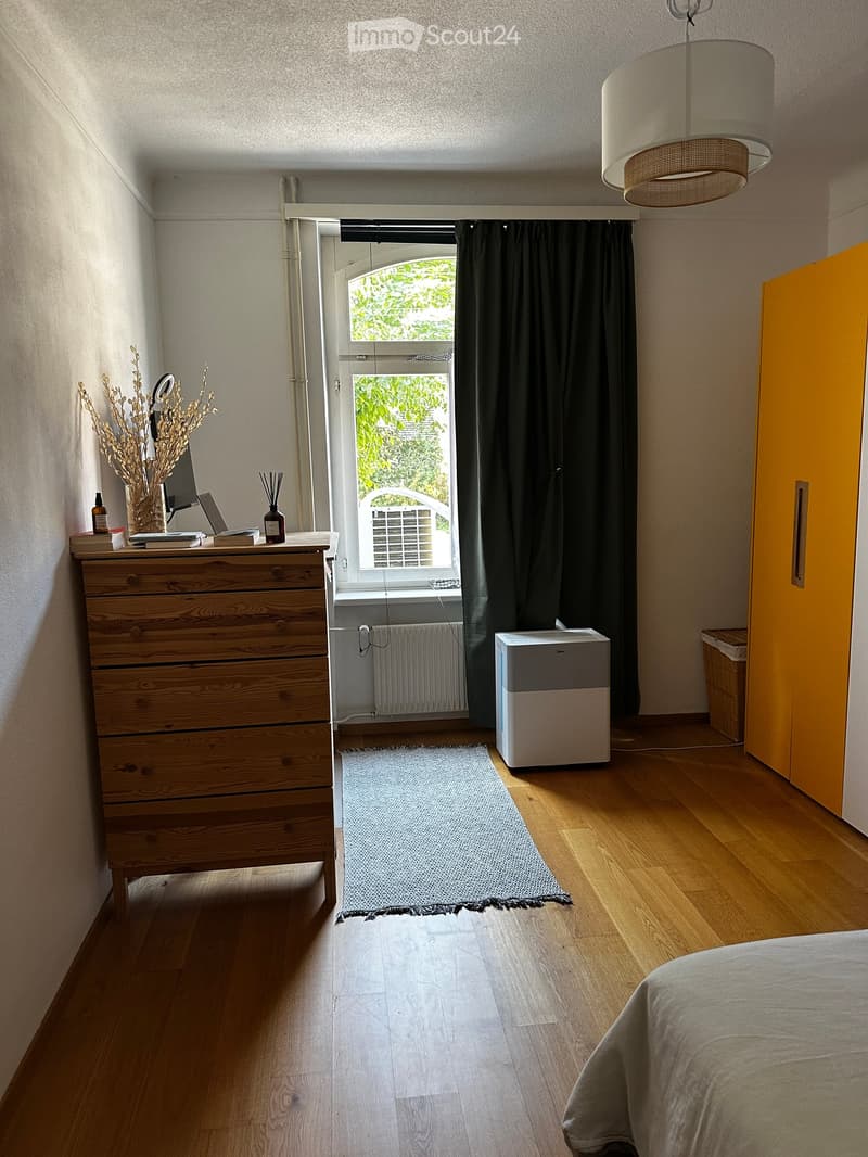 Cozy bedroom with wooden flooring, a bed with a grey duvet, a chest of drawers with a laptop and decorative items, a window with dark curtains, a radiator, an air conditioner, and a grey rug.
