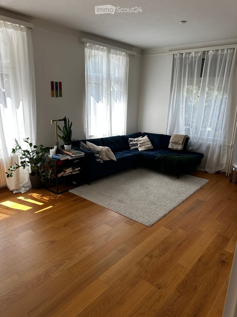 Modern living room with wooden flooring, large windows with white curtains, dark blue sectional sofa, coffee table with books and plants, and a white rug.