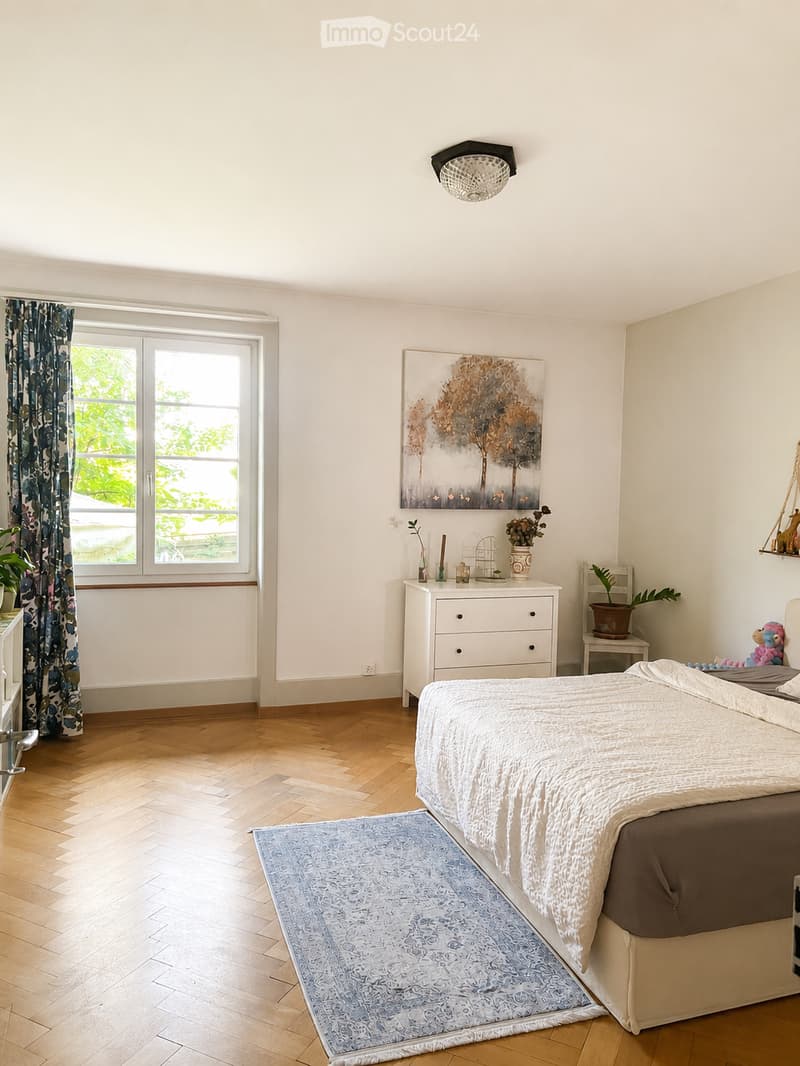 Bright bedroom with wooden flooring, large window with curtains, bed with white and brown bedding, nightstand, drawer, painting, rug, and small table with plants.