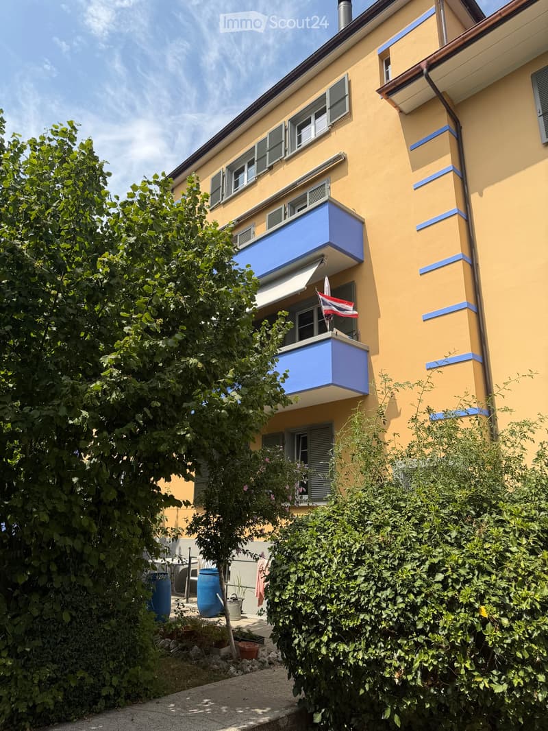 A yellow multi-story building with blue balconies and shutters, surrounded by greenery and outdoor furniture.