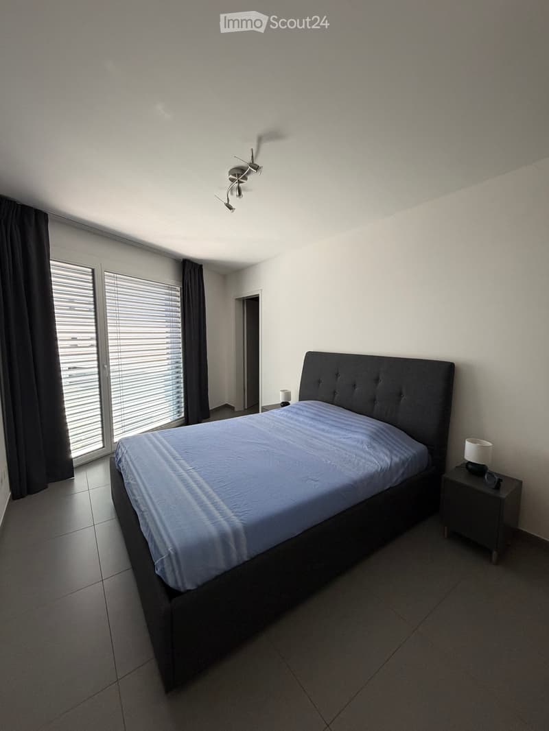 Modern bedroom with a large bed, nightstands, and lamps. Large windows with blinds and dark curtains provide natural light.