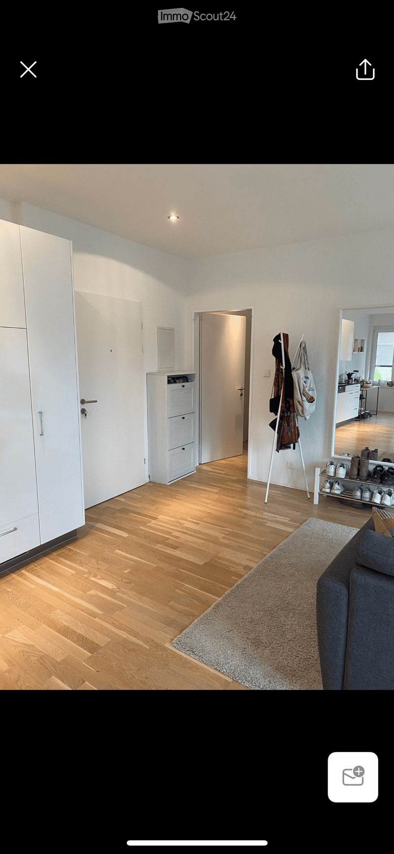 Bright living room with wooden flooring, a grey sofa, a large rug, a white wardrobe, a shoe rack, and a visible kitchen area with cabinets and appliances.