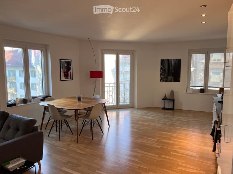Open-plan living and dining area with wooden flooring, large windows, and a balcony access. Features a sofa, dining table with chairs, a floor lamp, and decorative items.