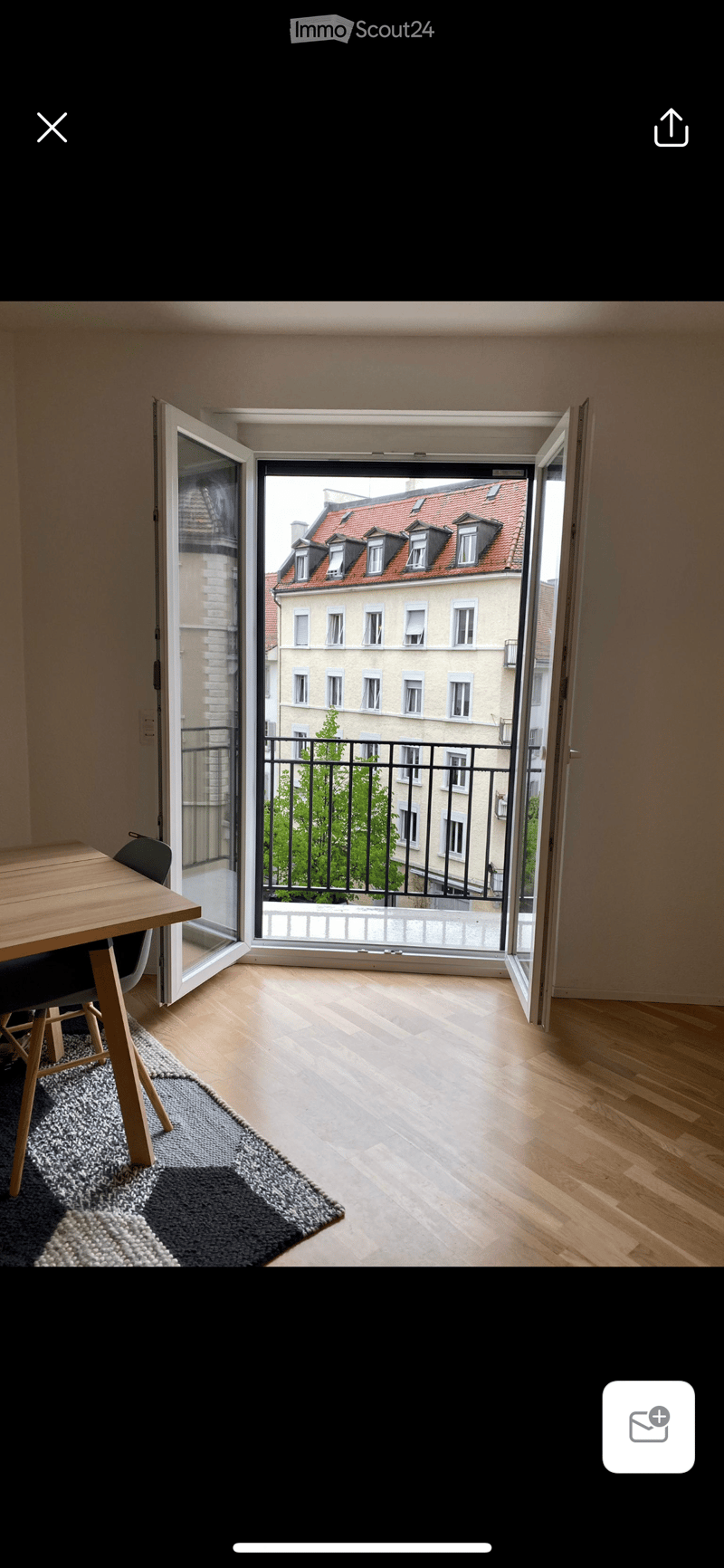 Bright room with wooden flooring, a small table and chair, and a rug near the door leading to a balcony with a view of a building.