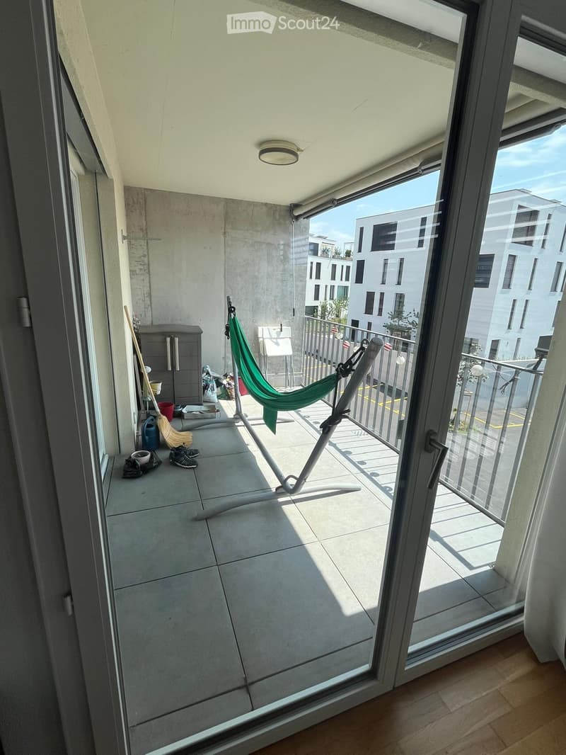 Balcony with tiled floor, glass sliding doors, a green hanging chair, a small table, and a view of a modern building.