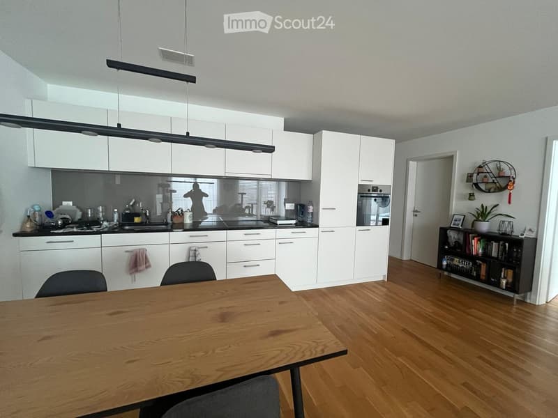 Modern kitchen with white cabinets, wooden dining table, and black countertop. Includes a sink, oven, and a small bookshelf with books and a plant.