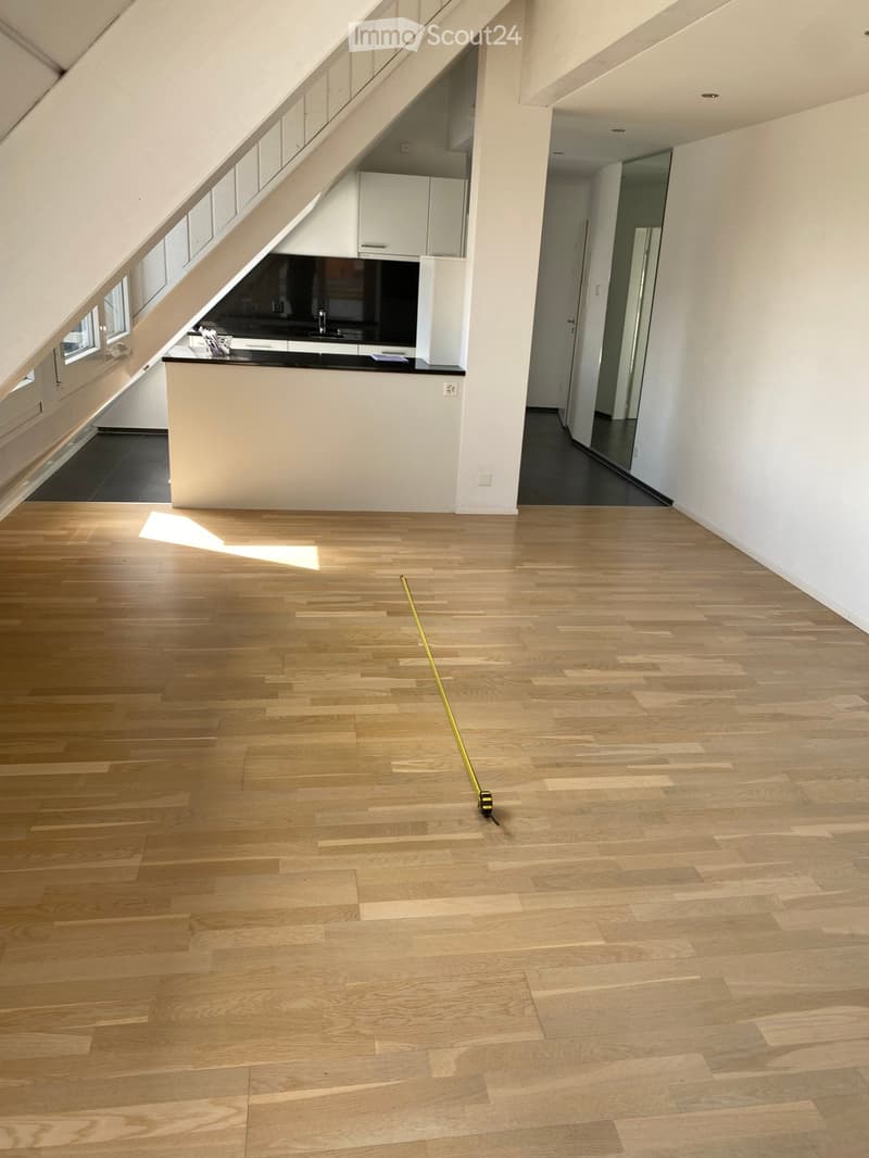 Empty room with wooden flooring, white walls, and a small kitchen area in the background.
