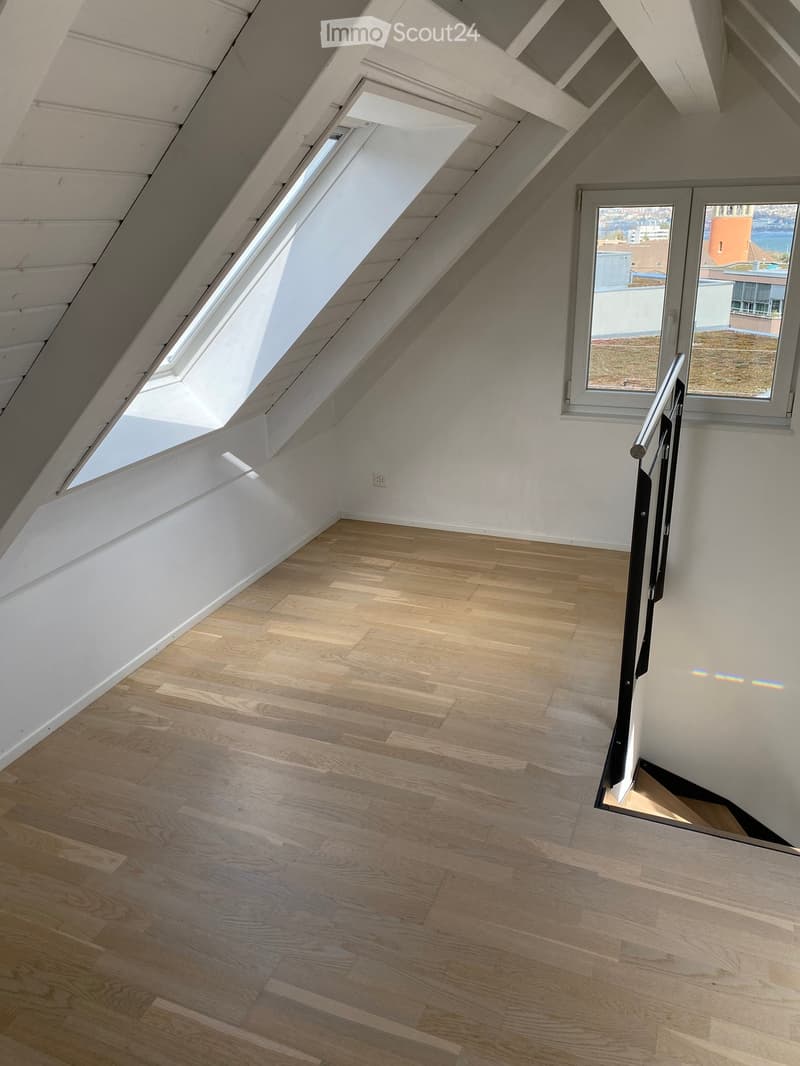 Empty attic room with wooden flooring, white walls, skylight windows, and a staircase leading downwards.