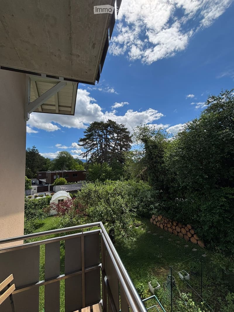 Balcony overlooking a lush, green garden with trees, bushes, and a stack of logs. The railing is metal, and the sky is partly cloudy.