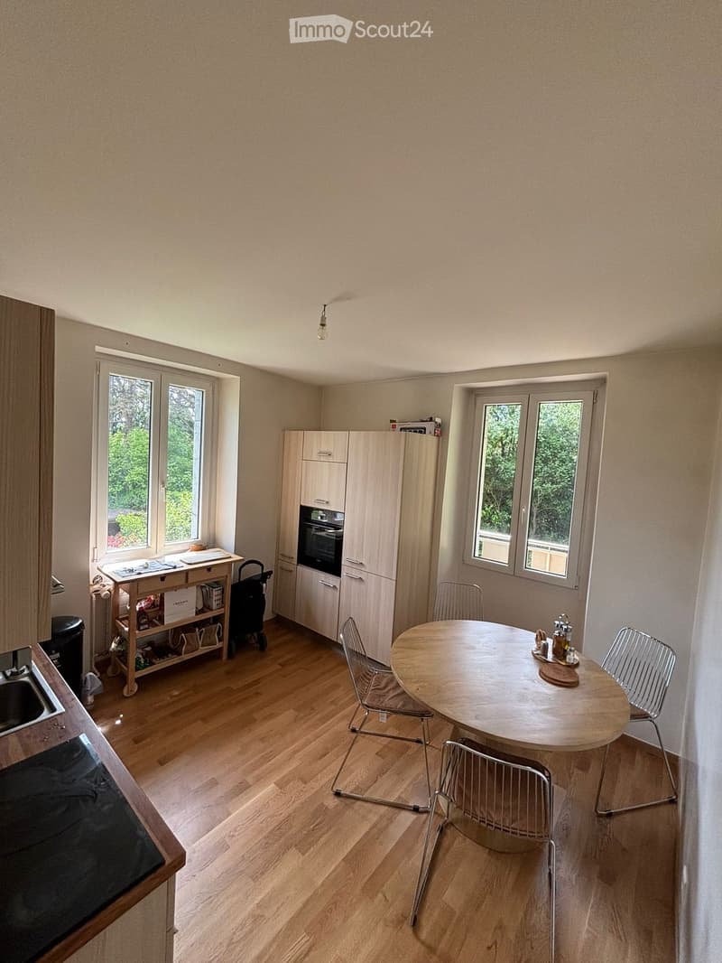 Bright kitchen with wooden flooring, white cabinets, a round dining table with four chairs, and a sink area with a window overlooking greenery.