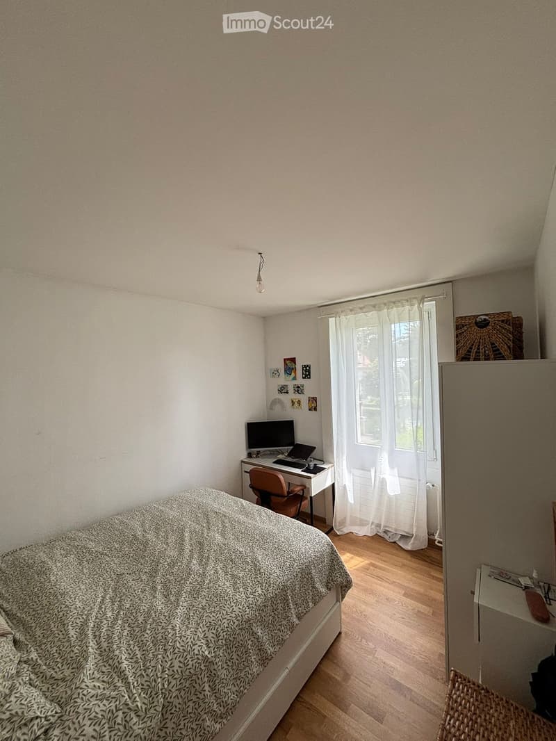 Cozy bedroom with a bed, desk, chair, laptop, computer monitor, and a window with white curtains. The room features light-colored walls and wooden flooring.