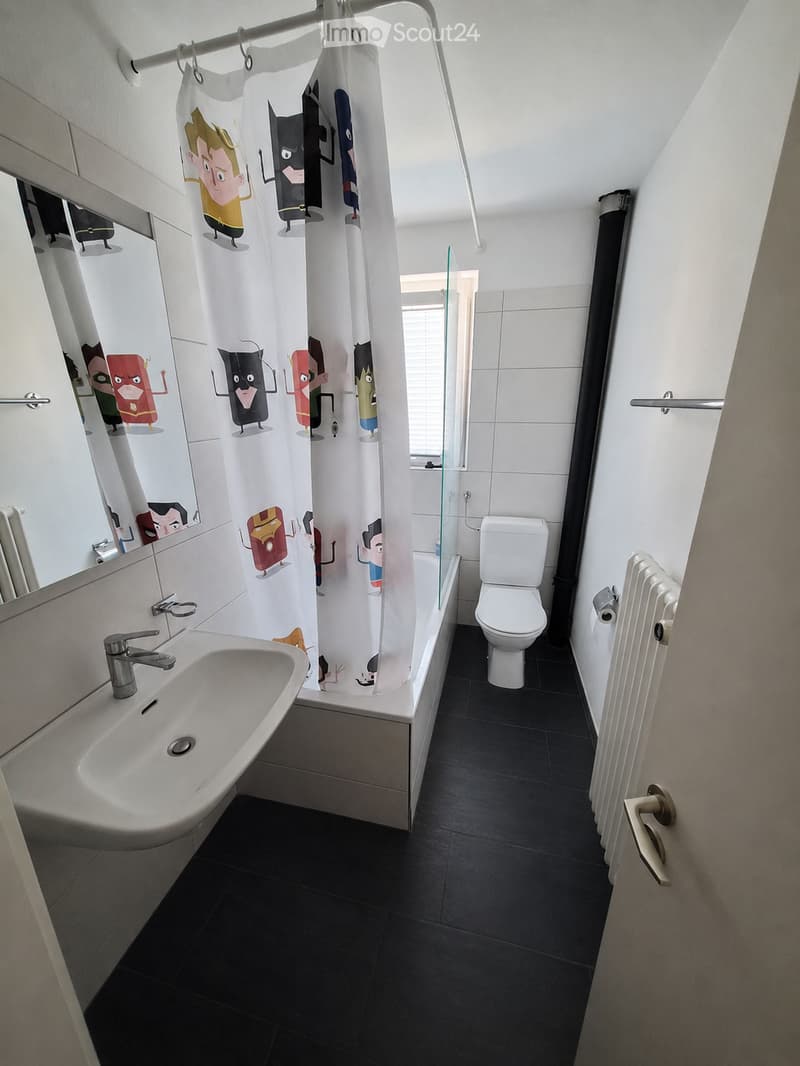 Modern bathroom with black tiled floor, white walls, sink, toilet, bathtub, radiator, and a colorful cartoon-themed shower curtain.